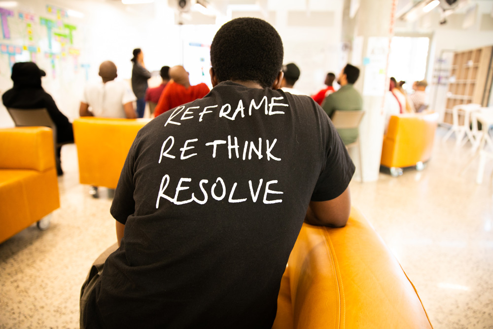 A man wearing a t-shirt at a d-school Afrika workshop, with the words &ldquo;Reframe, Rethink, Resolve&rdquo; written on the back.